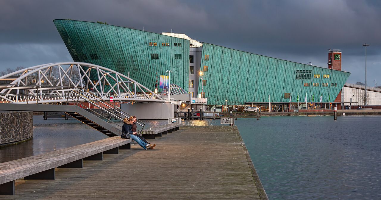 The copper-green NEMO Science Museum overlooks Amsterdam’s Oosterdok, with a pedestrian bridge and waterfront in the foreground.