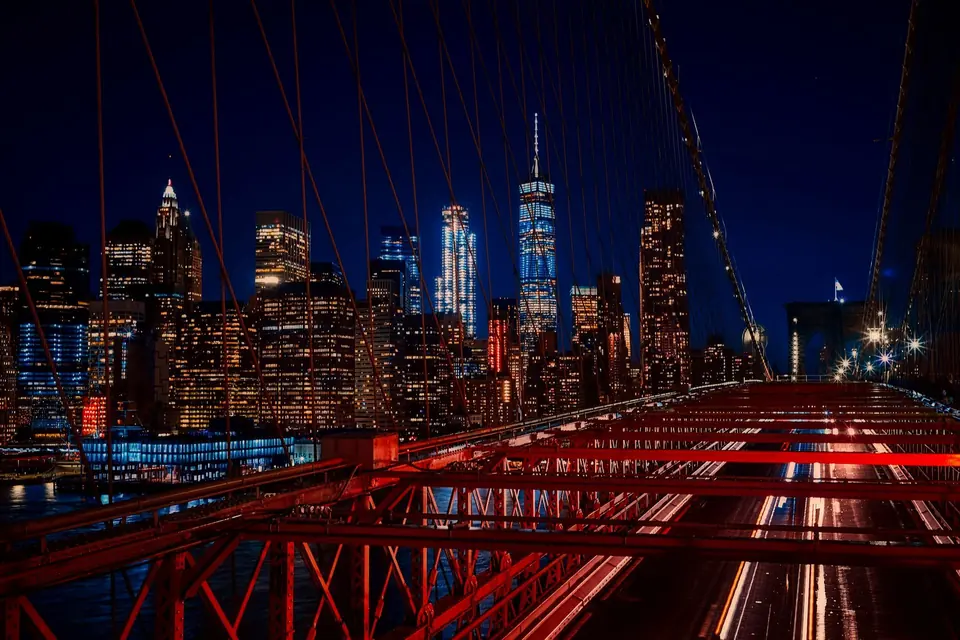 Brooklyn Bridge cables and roadway framing the illuminated Manhattan skyline at night.